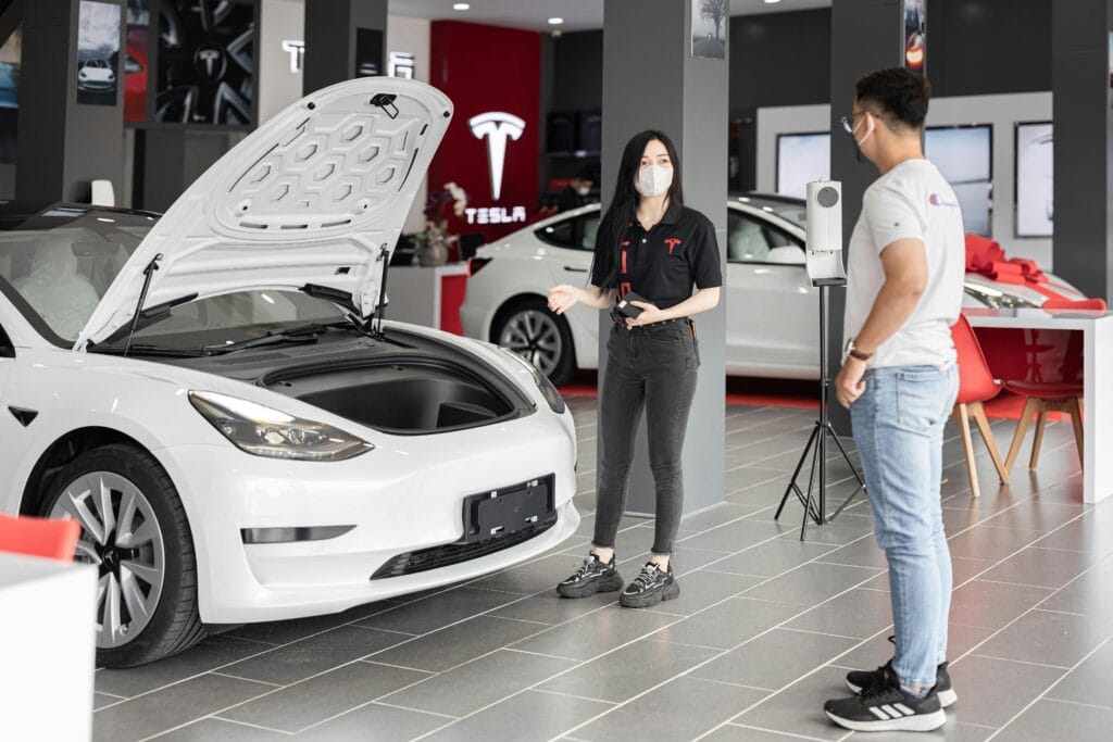 Tesla medarbejder forklarer funktioner i en hvid Tesla Model 3 med åben frunk til en kunde i en Tesla-butik.