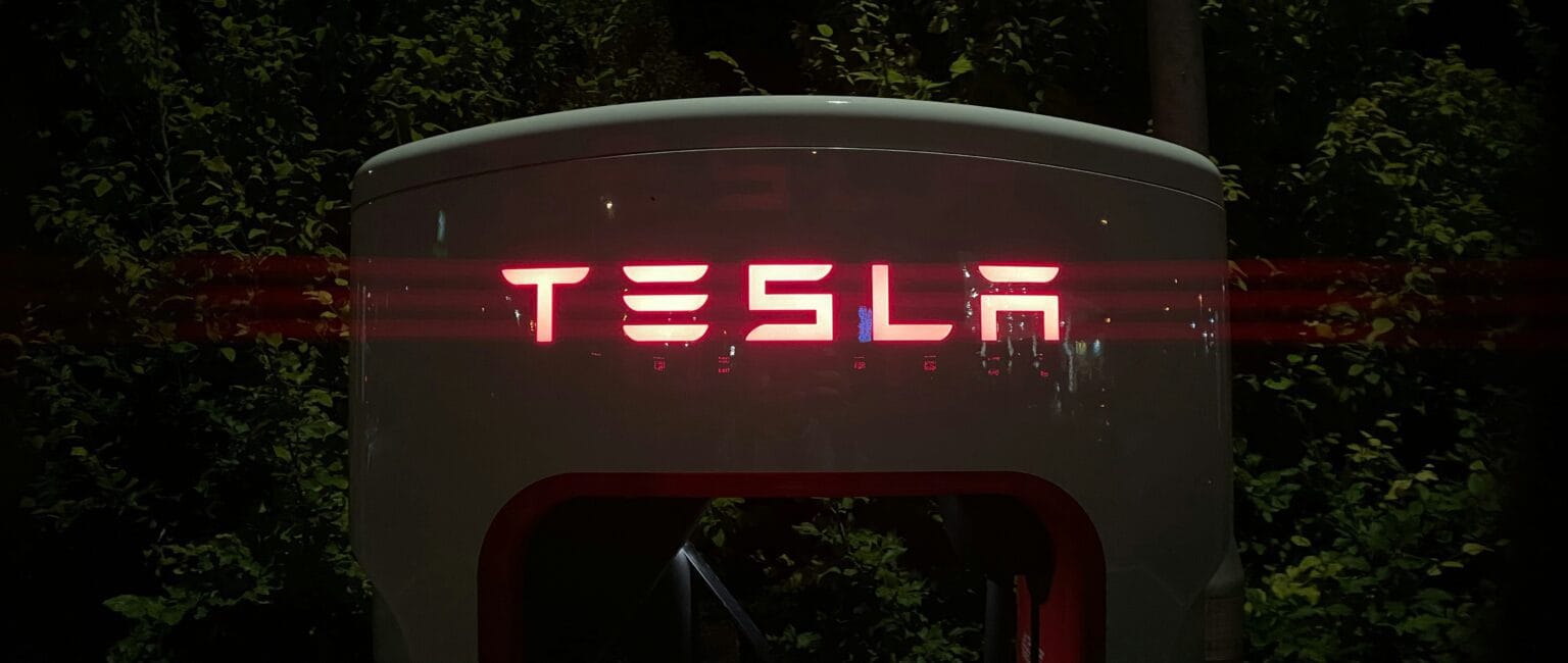 Tesla logo oplyst i rødt på en ladestation om natten omgivet af mørk vegetation.