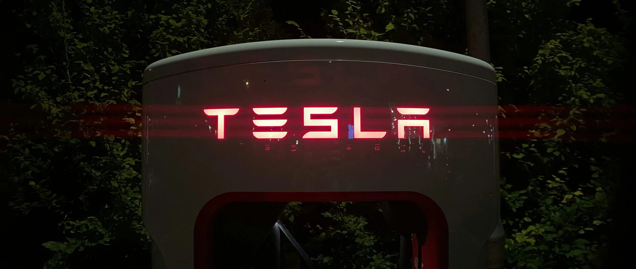 Tesla logo oplyst i rødt på en ladestation om natten omgivet af mørk vegetation.