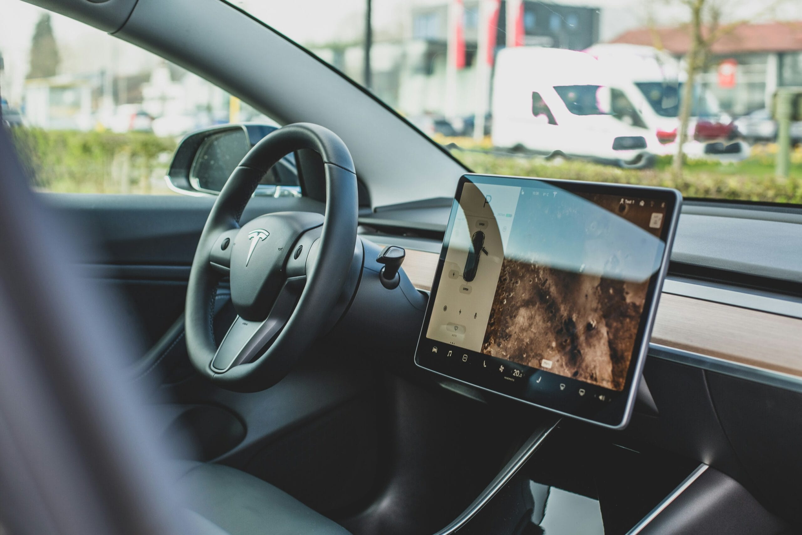 Interiør af en Tesla med rattet og den centrale touchskærm synlig, omringet af moderne design og en udsigt til biler udenfor.