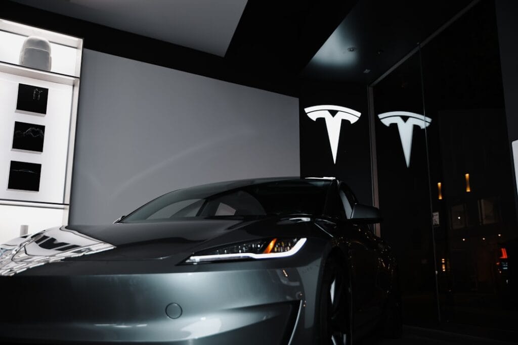 En grå Tesla-bil udstillet i et moderne showroom med Tesla-logoer oplyst i baggrunden.