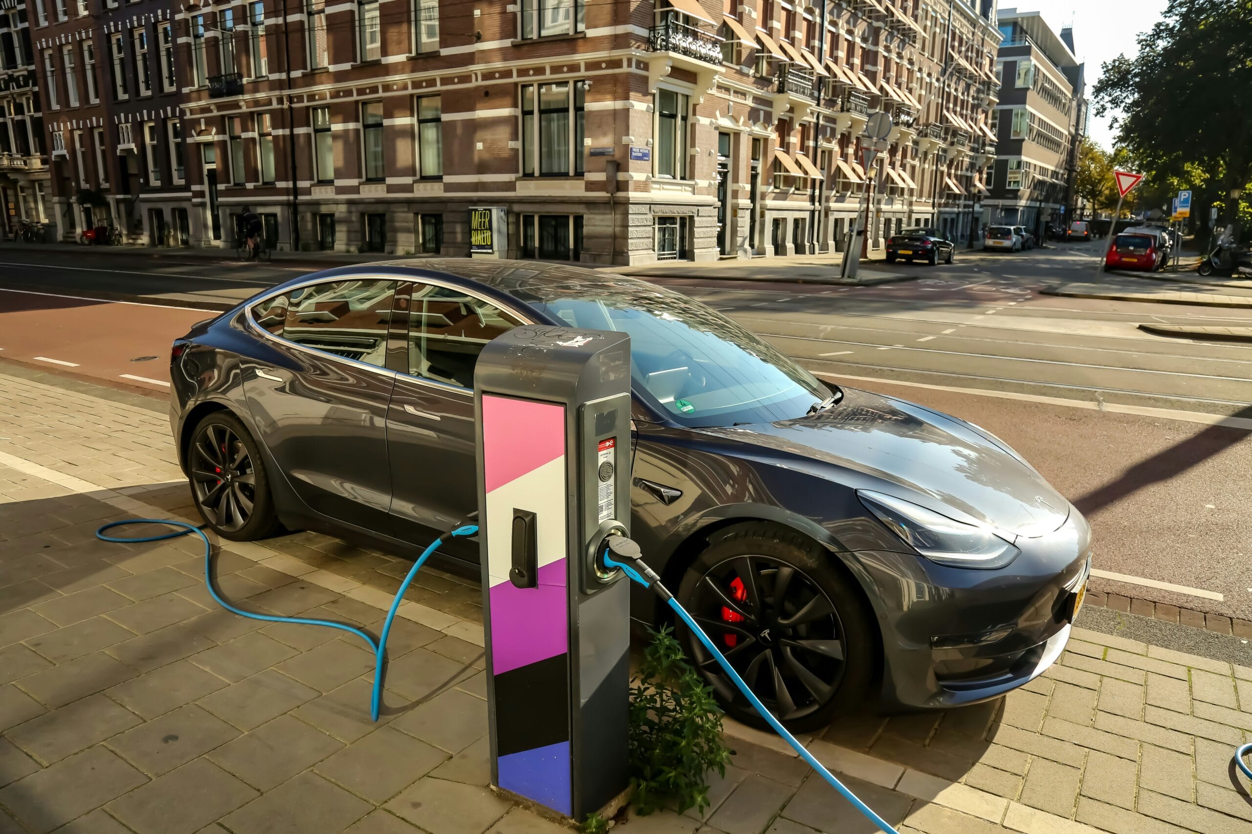 Tesla Model 3 til opladning ved en farverig ladestation i en urban gade med klassisk arkitektur.