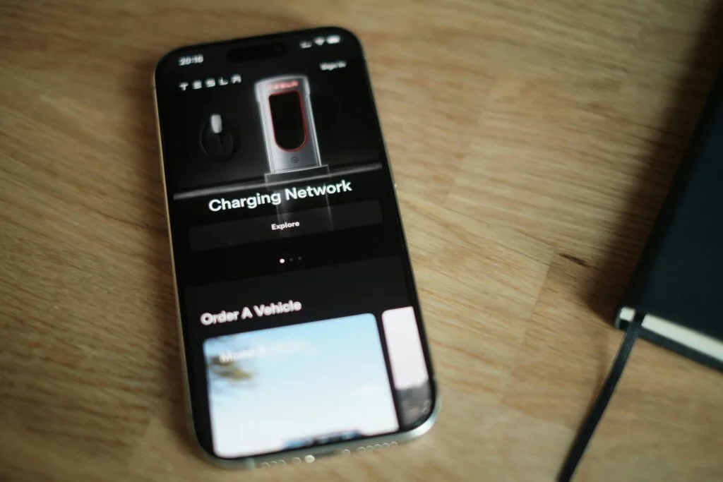 Smartphone med Tesla-app åben, der viser opladningsnetværk og køretøjsbestilling, placeret på en træbordsoverflade.