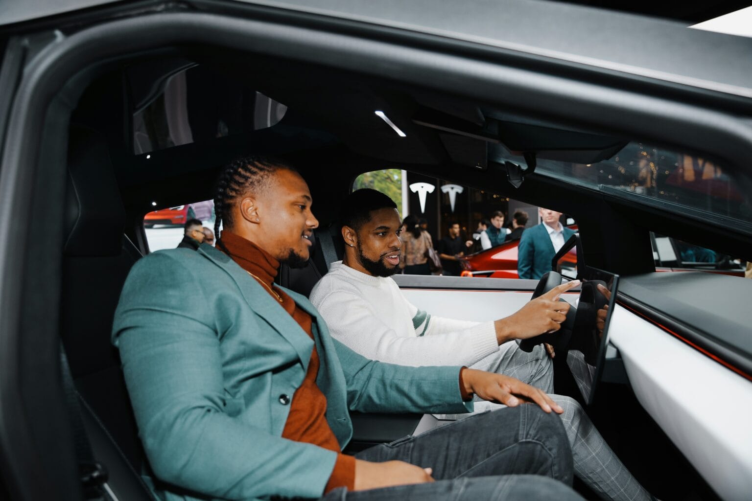 To mænd sidder i en Tesla og udforsker bilens interiør og teknologi under en udstilling med andre besøgende i baggrunden.