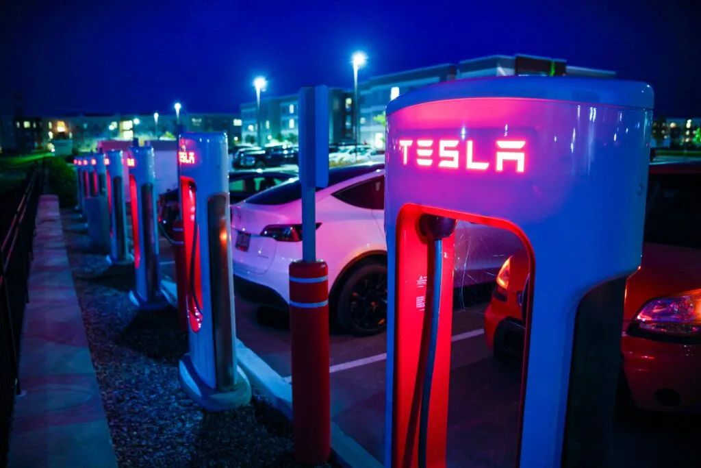 Tesla Supercharger-station oplyst i mørket med flere elbiler parkeret omkring.