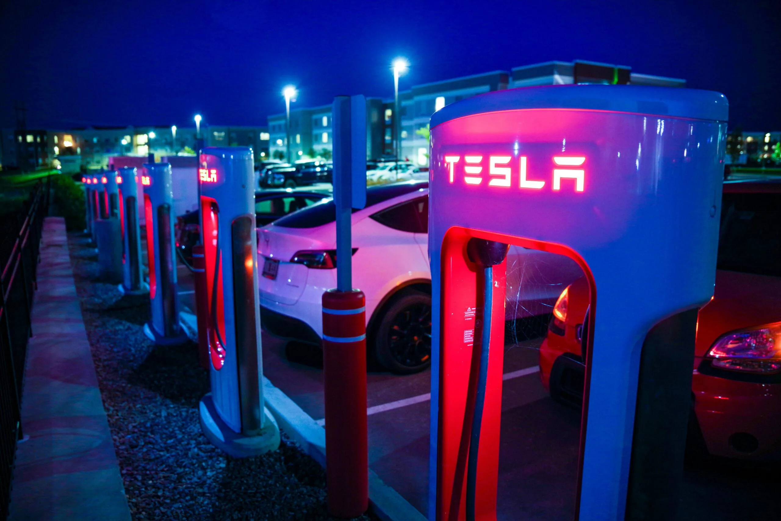 Tesla Supercharger-station oplyst i mørket med flere elbiler parkeret omkring.