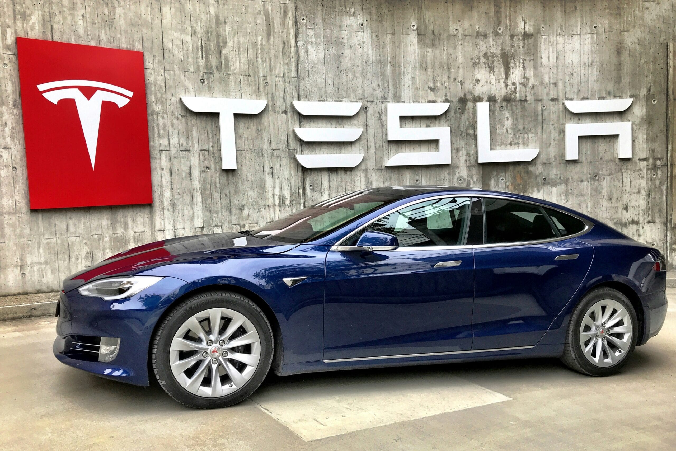 Blå Tesla Model S parkeret foran Tesla-logo på betonvæg