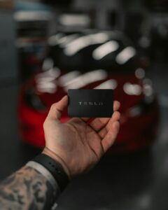 Hånd der holder et Tesla-kort foran en rød elbil, fokus på teknologi og innovation.