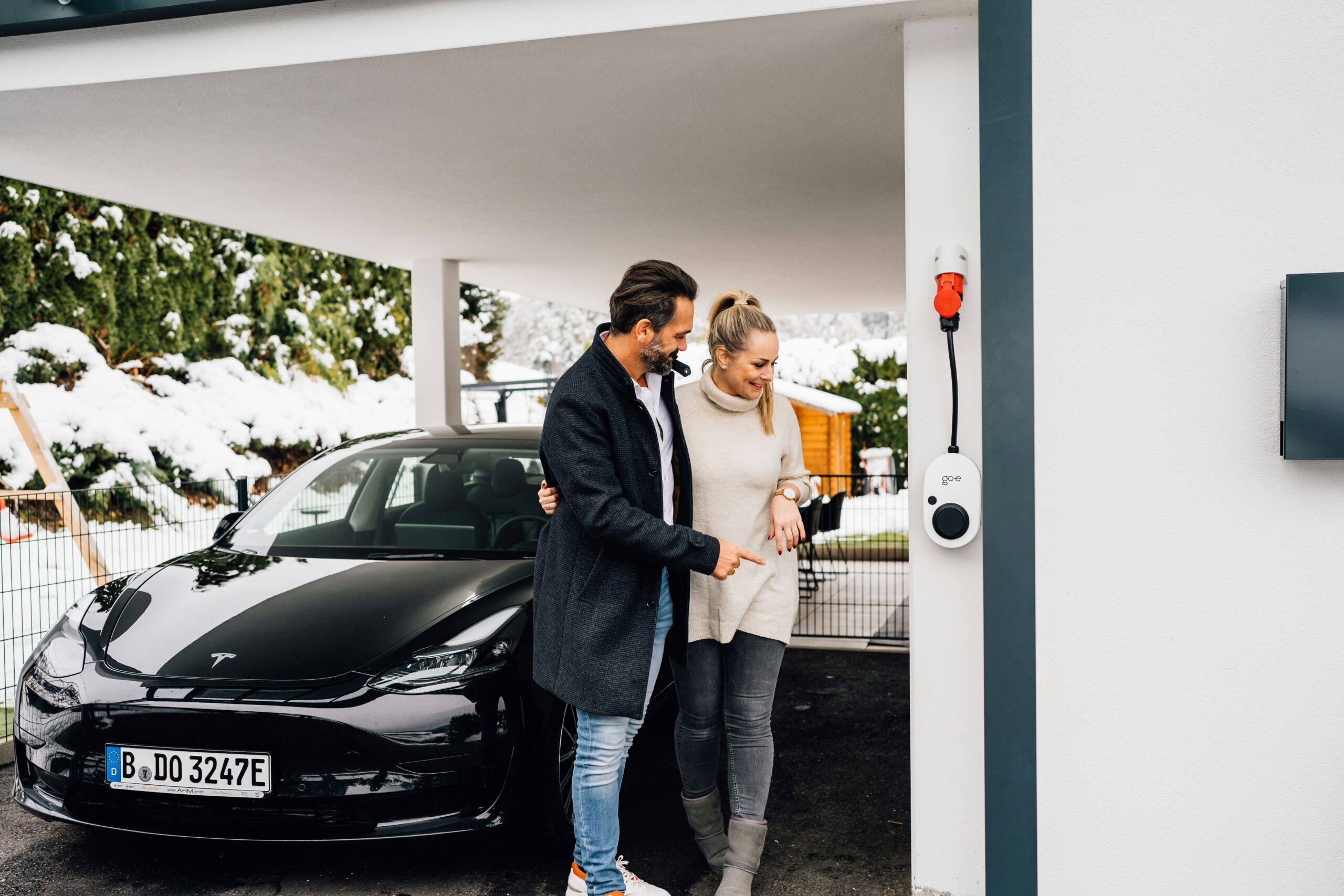 Par står ved en Tesla Model 3, mens de kigger på et ladepunkt hjemme i carporten.