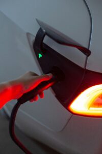 Hånd, der tilslutter en oplader til en Tesla elbil ved ladekontakt, med bilens lygter oplyst i baggrunden