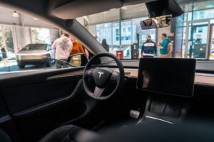 Interiør af Tesla med rat og stor skærm, set fra førersædet i et showroom med mennesker i baggrunden.
