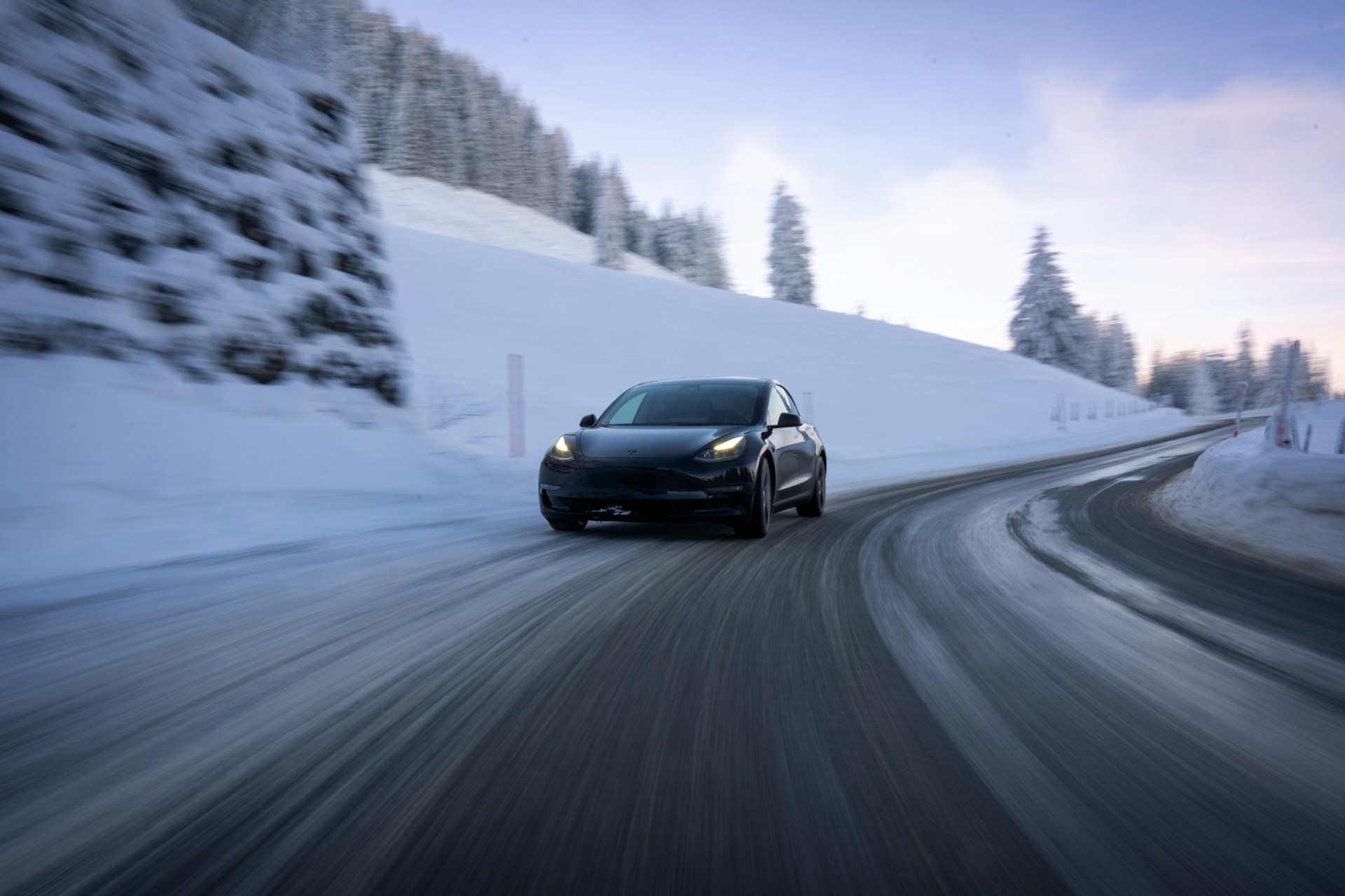 Mørk Tesla Model 3 kører på en ryddet bjergvej med sne i vejkanten