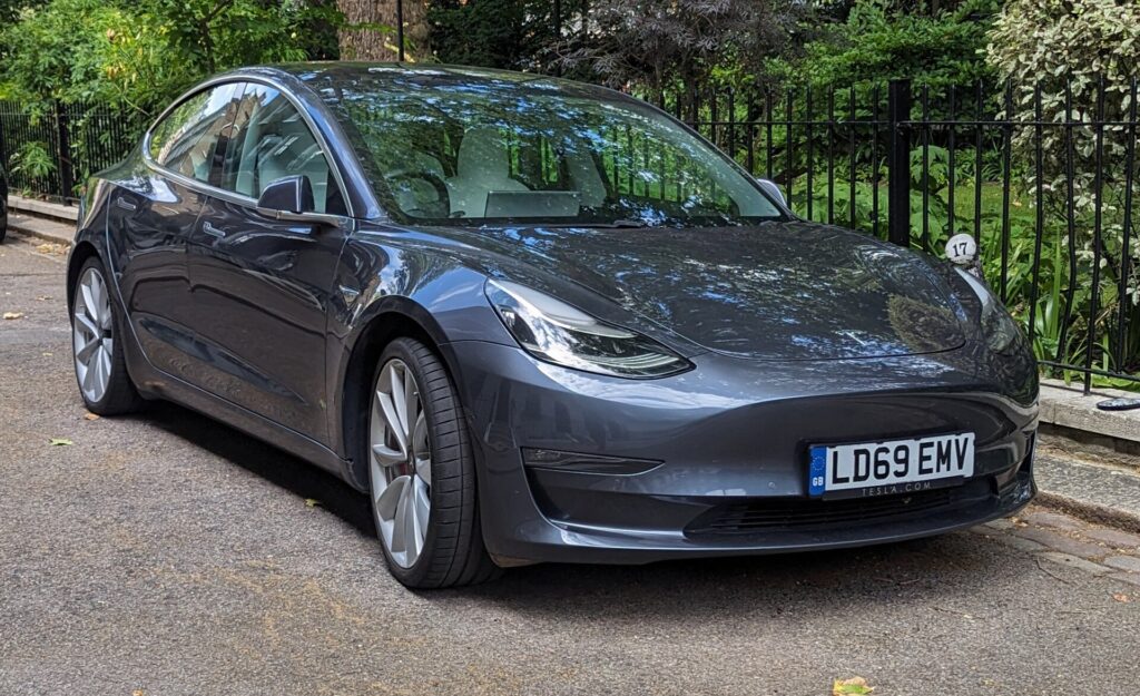 Grå Tesla Model 3 fra 2019 parkeret ved et fortov foran et jerngelænder