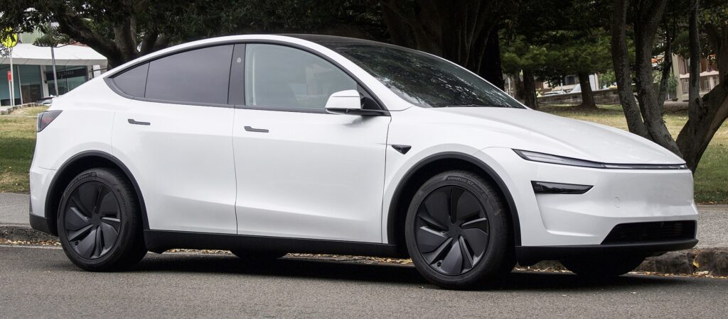 Hvid Tesla Model Y af den nye generation med baghjulstræk set skråt forfra, parkeret ved en vej med træer
