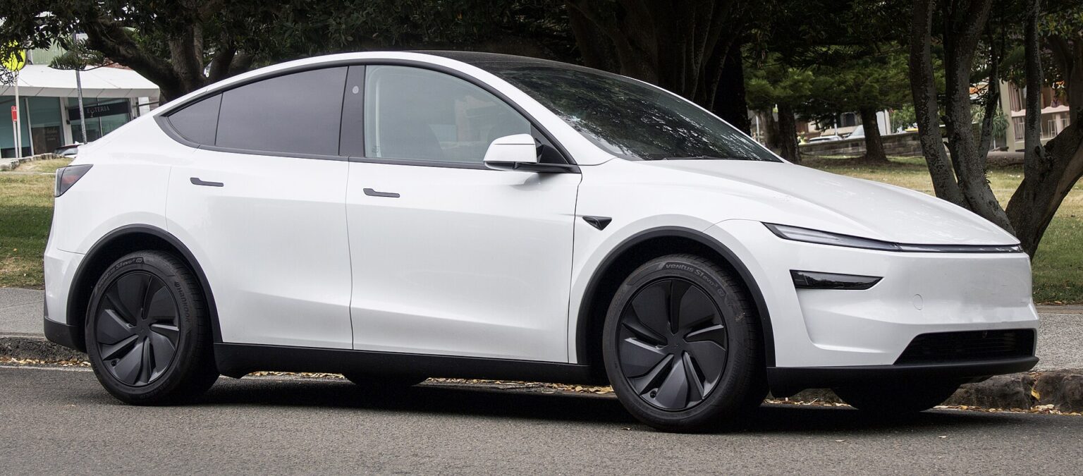 Hvid Tesla Model Y af den nye generation med baghjulstræk set skråt forfra, parkeret ved en vej med træer