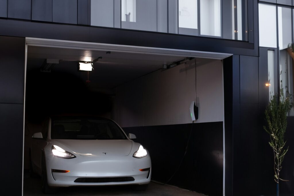 Tesla Model 3 med tændte forlygter i en åben garage, hvor en Wall Connector sidder på bagvæggen